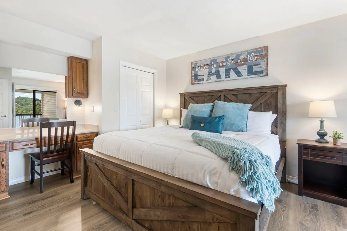 A cozy bedroom with a bed adorned with decorative pillows, a wooden headboard, lake-inspired wall art, and a desk with a chair. Natural light streams in, highlighting the room's inviting atmosphere.