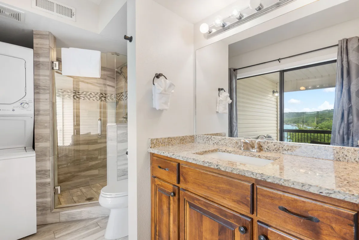 A contemporary bathroom featuring a granite countertop, glass shower, built-in laundry unit, and a large window with views outside. The setting is bright, modern, and highly functional.