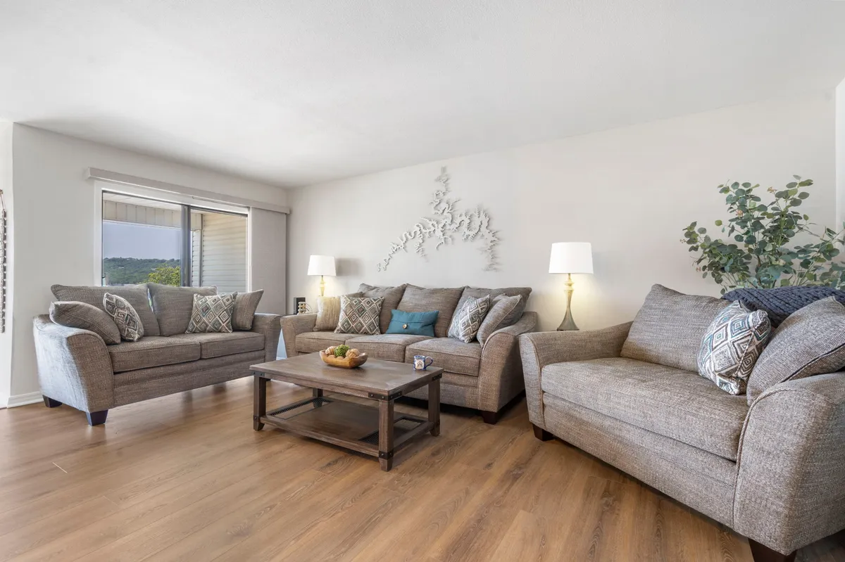 The image showcases a bright, welcoming living room with plush, comfortable sofas arranged around a modern coffee table. Tasteful decorative wall art and lush indoor plants add warmth and character, while the layout invites guests to relax, chat, or gather for a movie or game night. The space is styled for comfort and togetherness, reflecting the spirit of a lakeside retreat.