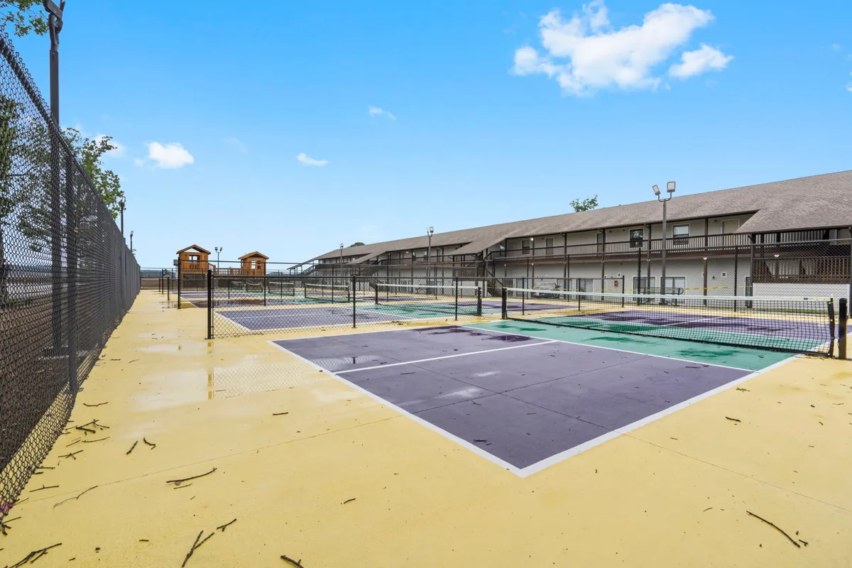 This exterior image captures vibrant tennis courts surrounded by trees and adjacent playground structures, with families and children engaging in active play and sports. The scene is lively, suggesting a welcoming community environment and ample outdoor recreational options.
