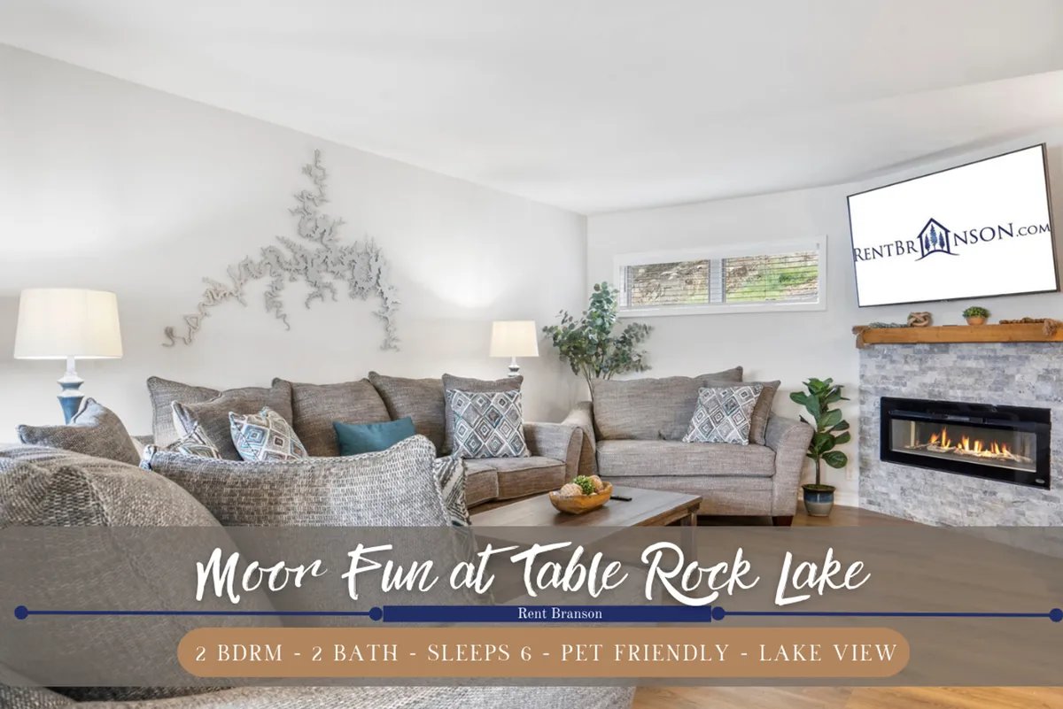 A warm, inviting living room featuring plush, comfortable sofas arranged around a classic fireplace, with a large television mounted above. Expansive windows reveal glimpses of the lake beyond, while decorative wall art and soft lighting add to the welcoming ambiance. The space seamlessly connects to the main living area, ideal for family gatherings, movie nights, and relaxing after a day by Table Rock Lake.