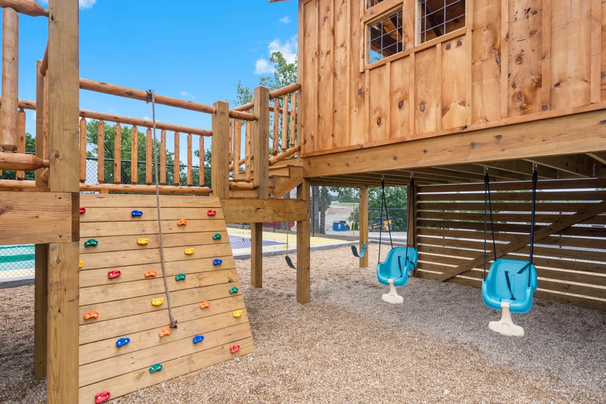 A vibrant outdoor playground surrounded by lush greenery, featuring a colorful play structure with a rock climbing wall and multiple swings. Children are actively climbing, swinging, and playing, creating a joyful, energetic atmosphere.