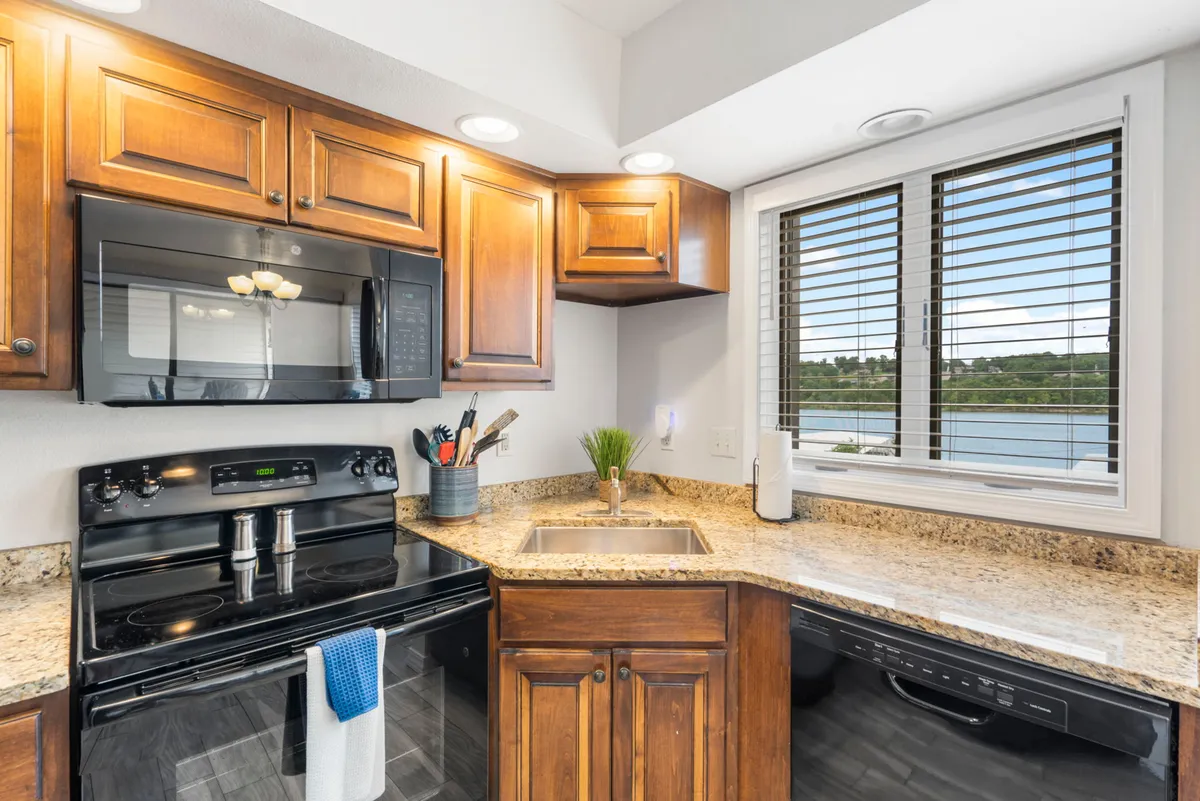 A view of a warmly lit kitchen featuring granite countertops, dark wooden cabinets, and modern stainless steel appliances. A window over the counter lets in natural light, and the open layout invites gathering and conversation.