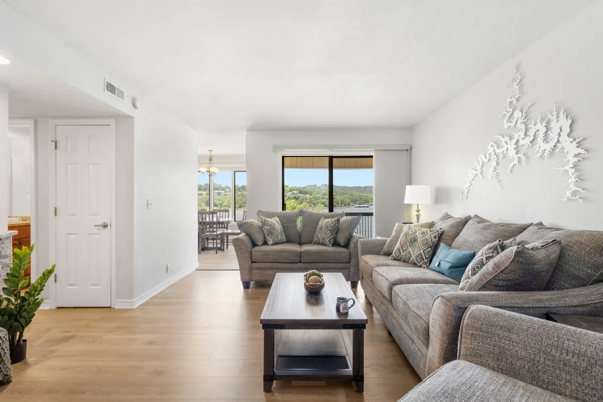 A cozy living area with soft, comfortable sofas facing a coffee table adorned with a simple plant. Decorative wall art adds personality, while a large window frames a breathtaking sunset view over the lake, flooding the room with warm, natural light. The space feels perfect for relaxing, reading, or intimate conversations.