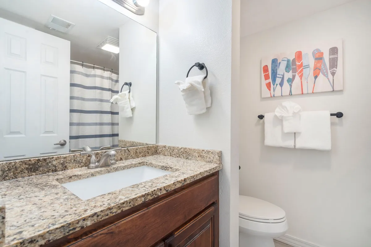 A well-lit bathroom with a granite countertop, large mirror, colorful artwork, and neatly arranged towels. The ambiance is fresh, inviting, and ideal for self-care routines.
