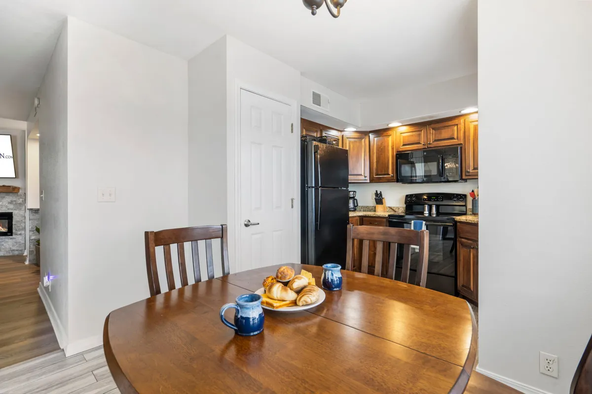 A dining table set for breakfast, complete with pastries, coffee mugs, and kitchen appliances visible in the background. The scene feels inviting and intimate, suggesting shared meals and leisurely mornings.