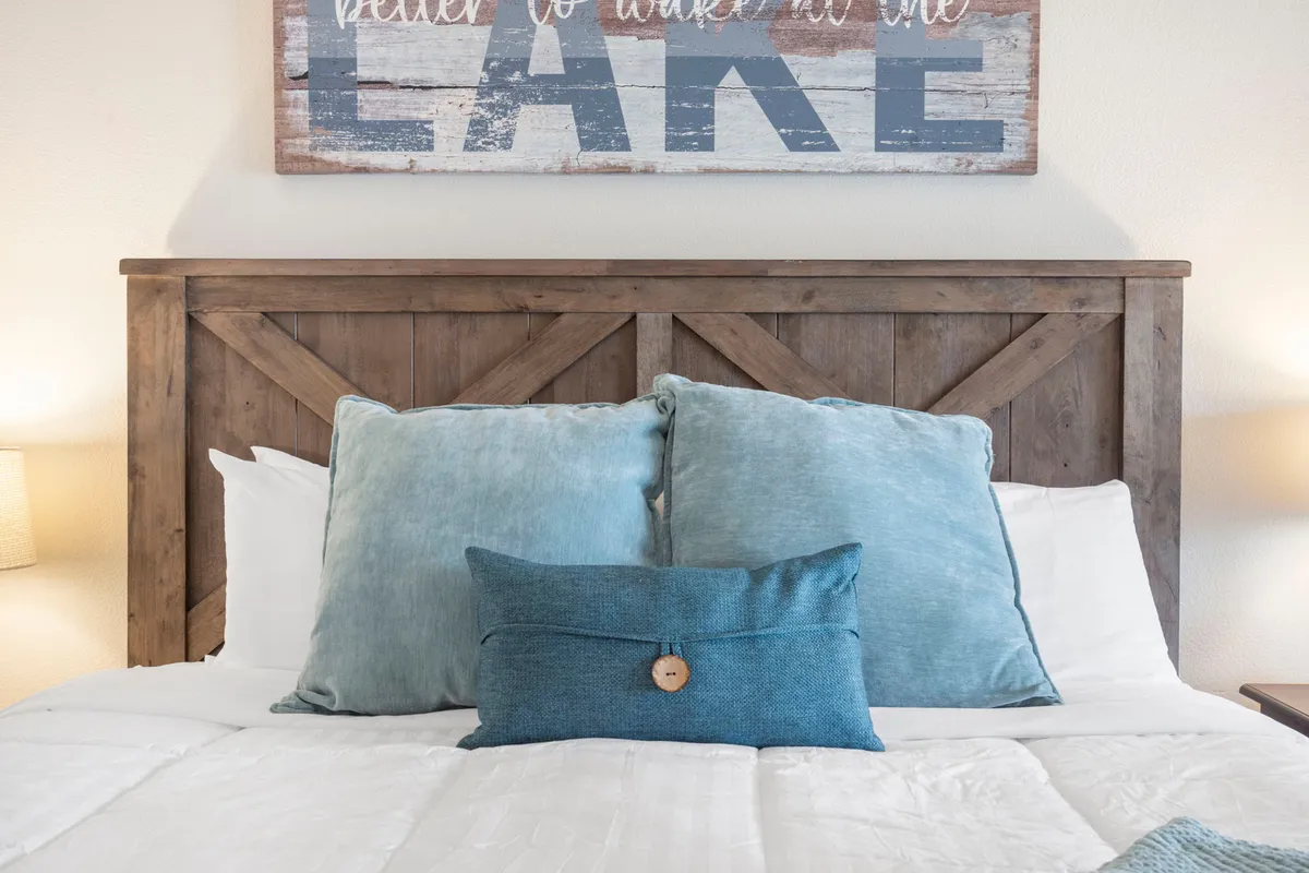 A king bedroom featuring a wooden headboard, decorative pillows, and lake-themed wall art. The space is softly lit and exudes a tranquil, restful ambiance perfect for unwinding after a day of activities.