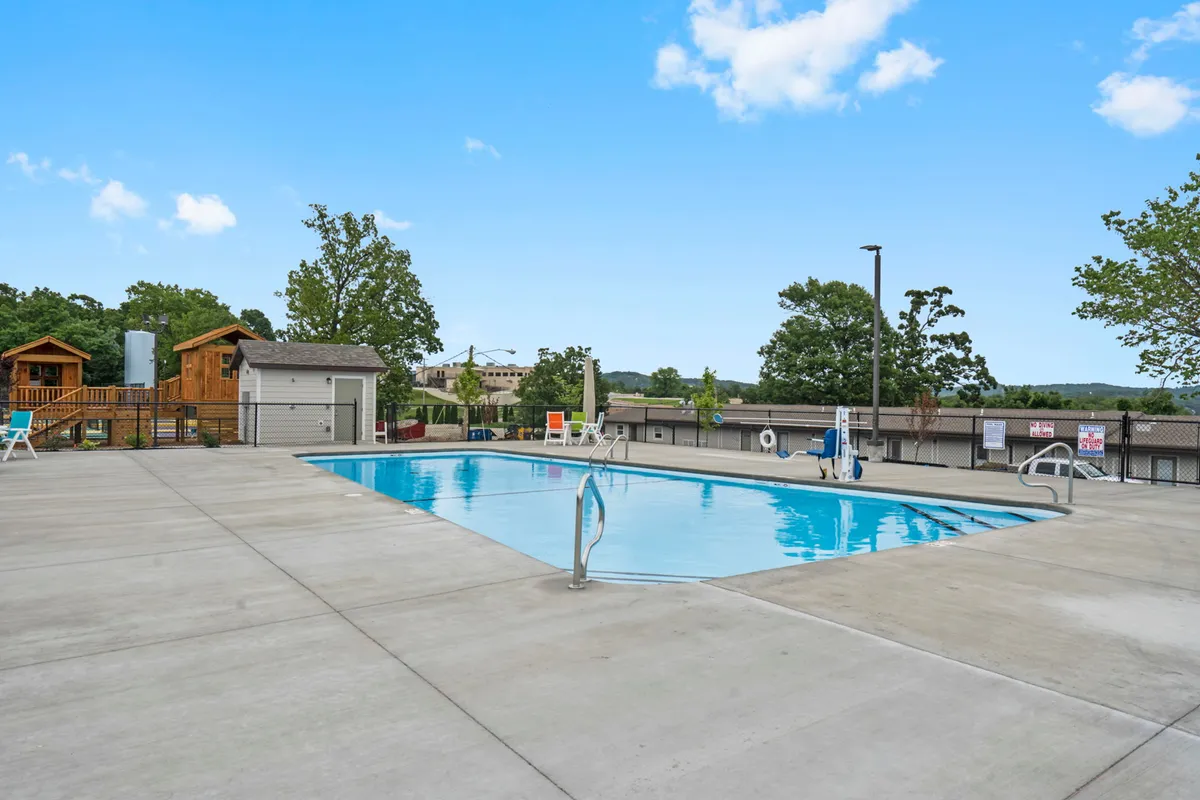 An inviting outdoor pool scene featuring a sparkling pool surrounded by deck chairs and lush trees, with a building structure nearby. The setting suggests a vibrant atmosphere perfect for swimming, sunbathing, and socializing, suitable for both families and groups.